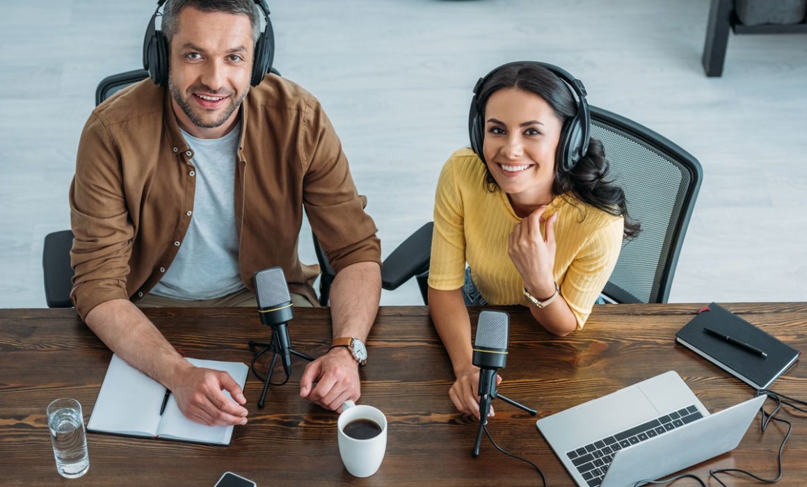 Smiling male and female podcast hosts wearing headphones, with mics at a table.