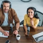 Smiling male and female podcast hosts wearing headphones, with mics at a table.